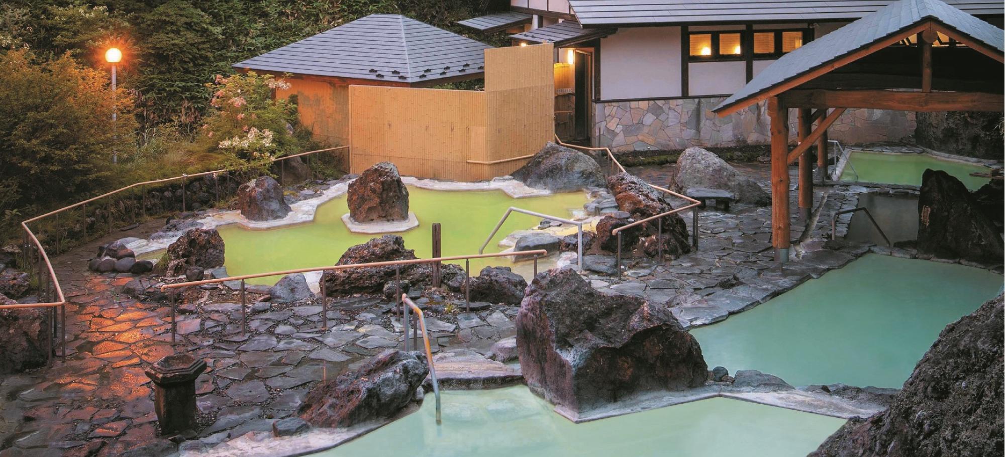 万座高原ホテル — 嬬恋村万座 · 岩を配した石庭露天風呂に白濁と緑色の湯船が並ぶ夕景