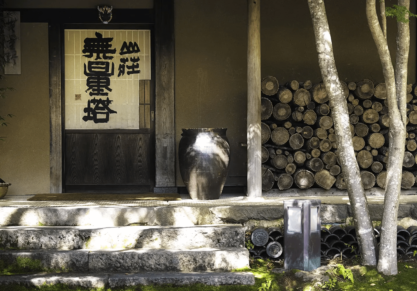 山荘 無量塔 — 由布院・川上 · 墨書きの屋号を掲げた玄関と薪が積まれた軒先